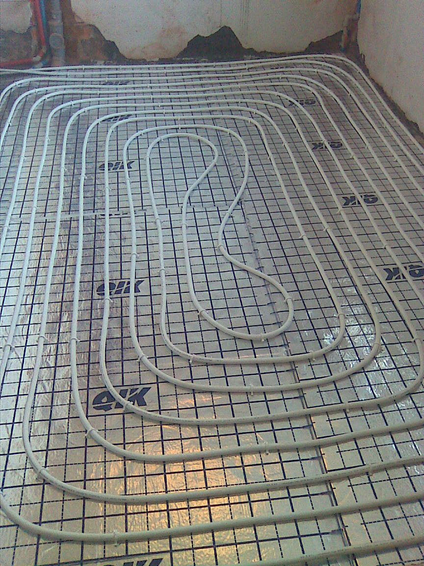 Białe rury ogrzewania podłogowego ułożone spiralnie na srebrnej macie z czarną siatką, widoczne fragmenty ściany w tle.